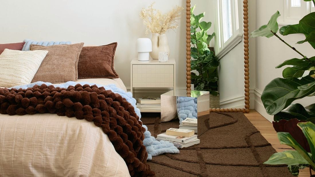 A bedroom with a mirror cube on a brown rug next to a bedside table with a lamp and vase, and a bed with brown and blue blankets. A tall, beaded floor mirror and a large green plant are also visible.