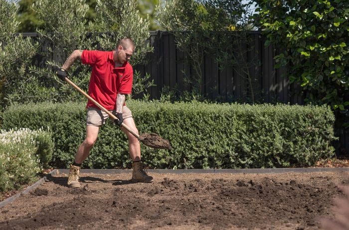 A Bunnings Team Member is using a shovel to dig the area to calculate the required amount of road base and paver sand for the project.