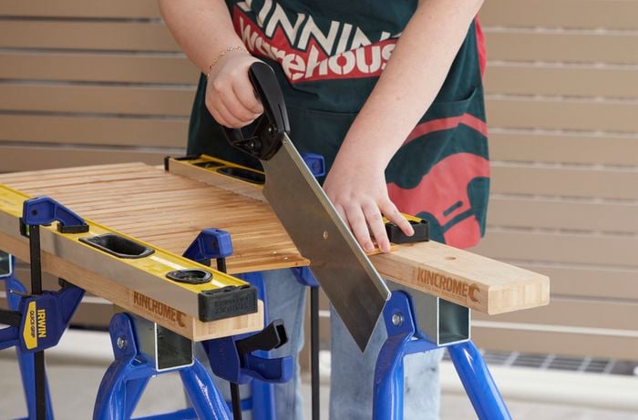 Person using handsaw to cut through mat, resting on sawhorses.