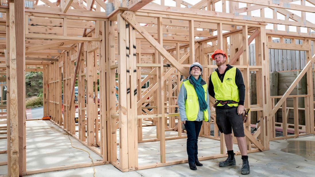 A woman and man, both wearing high-vis vests and hardhats, are looking up to inspect roofing trusses inside the frame of a house under construction.