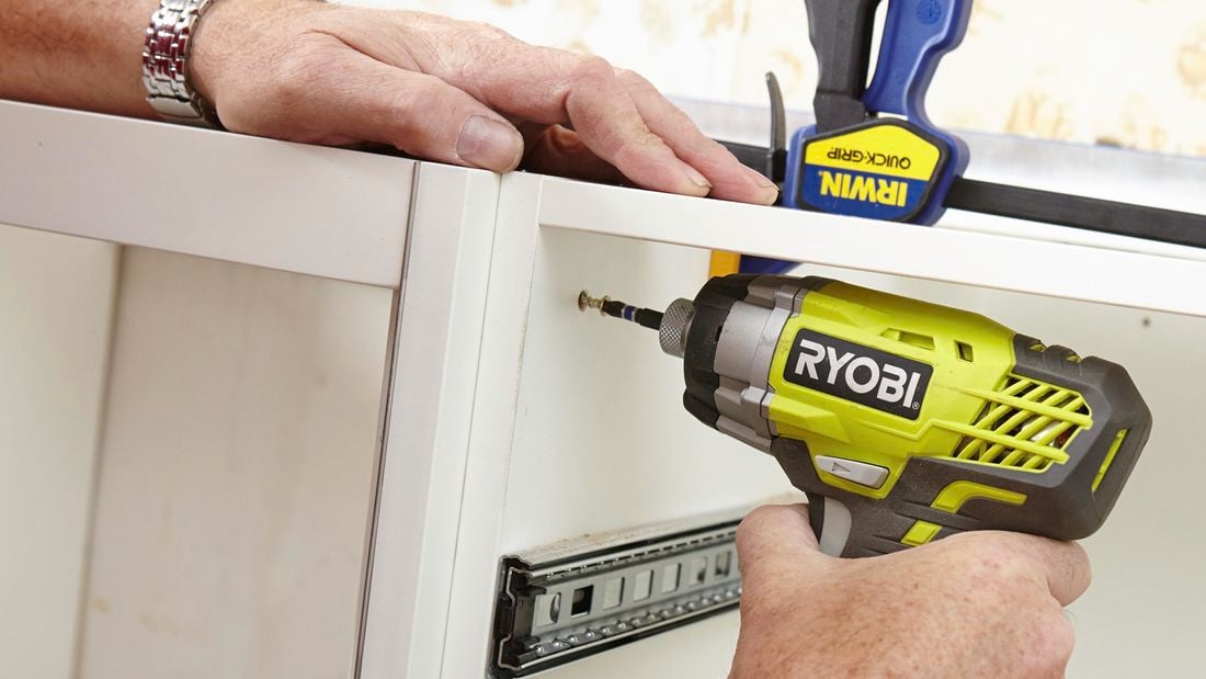 Two kitchen cabinets being screwed together with a power drill, while held together with clamps