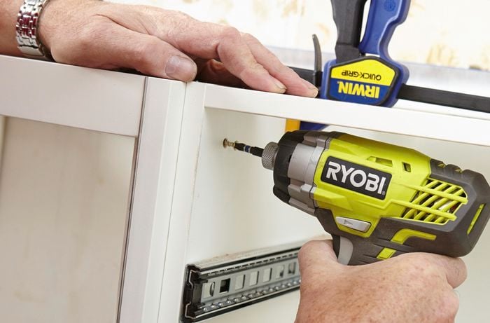 Two kitchen cabinets being screwed together with a power drill, while held together with clamps