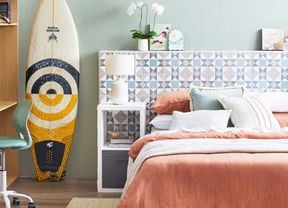  A light-coloured tiled D.I.Y. bedhead with green plants on top, behind a bed in a room with a surfboard, chair and rug