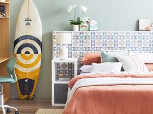  A light-coloured tiled D.I.Y. bedhead with green plants on top, behind a bed in a room with a surfboard, chair and rug