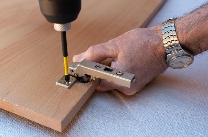 A person is using a drill to unscrew and remove a metal hinge attached to the doors and cabinetry.