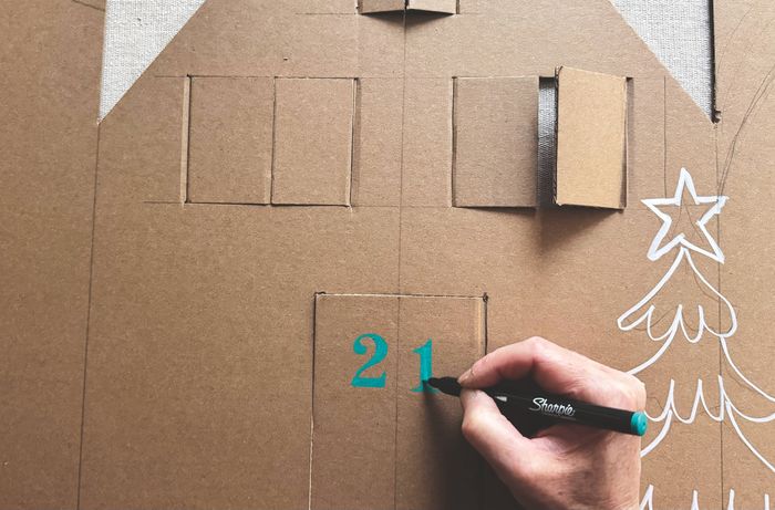 A hand using a marker to write numbers on a cardboard Advent calendar.