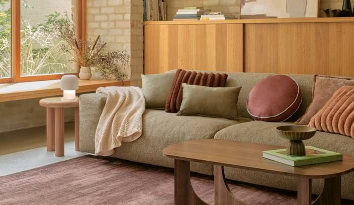 Minimal living room corner featuring a green upholstered lounge chair with a neutral cushion, a small wooden side table with a green lamp, a timber bookshelf, and a dark round rug on timber floors.