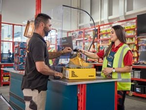 A male tradie holds up his Bunnings Trade PowerPass App to be scanned by a Bunnings Warehouse Red Shirt team member in store, with yellow DeWALT boots on top of a box on the counter.