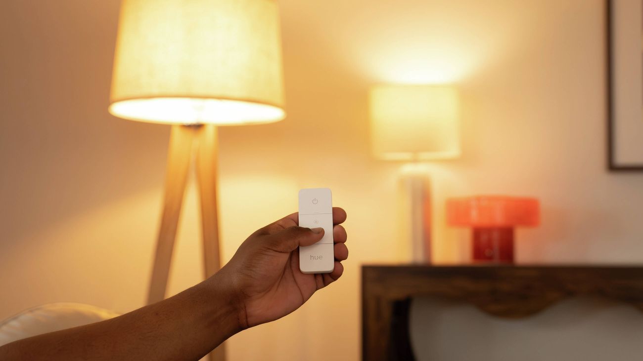 Hand holding a Philips Hue smart lighting remote control in a warmly lit living room with illuminated lamps in the background.