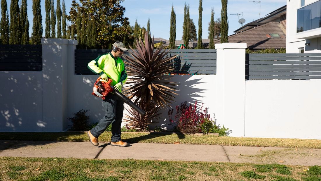 A landscaper wearing high‑visibility clothing uses a leaf blower to clear grass clippings and debris from a path beside a residential garden.