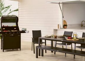  A black barbecue grill with its lid open, cooking onions, bell peppers and flatbreads on the griddle, set on a patio next to a dining table and chairs.