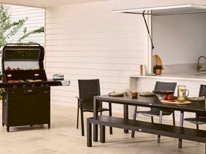  A black barbecue grill with its lid open, cooking onions, bell peppers and flatbreads on the griddle, set on a patio next to a dining table and chairs.