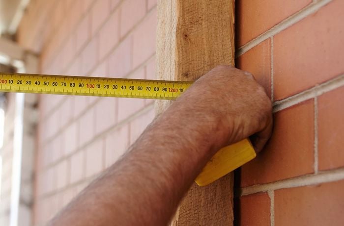 A person measuring distance from a timber post against a brick wall