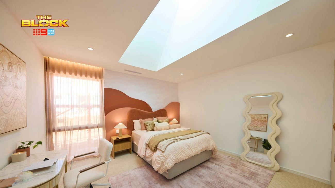 Cream bedroom with a queen bed, desk and mirror on a blush rug and a red and orange hill style bedhead.