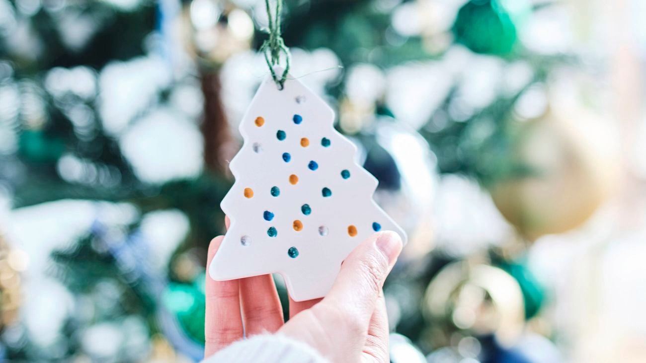 Close up image of a hand holding a white clay Christmas tree ornament with coloured dots, with a Christmas tree in the background.