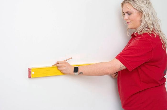 A Bunnings team member is using a spirit level and pencil to draw a horizontal line on the wall before painting a scalloped wall pattern.