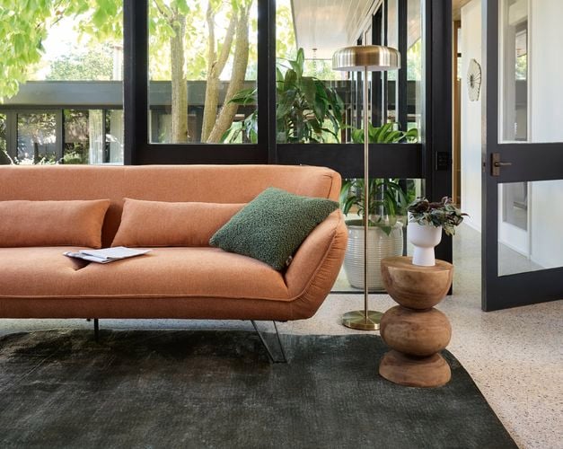 A lounge room with an orange couch with a green cushion on it, a light stand, a rug on carpet in it.