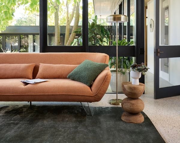 A lounge room with an orange couch with a green cushion on it, a light stand, a rug on carpet in it.