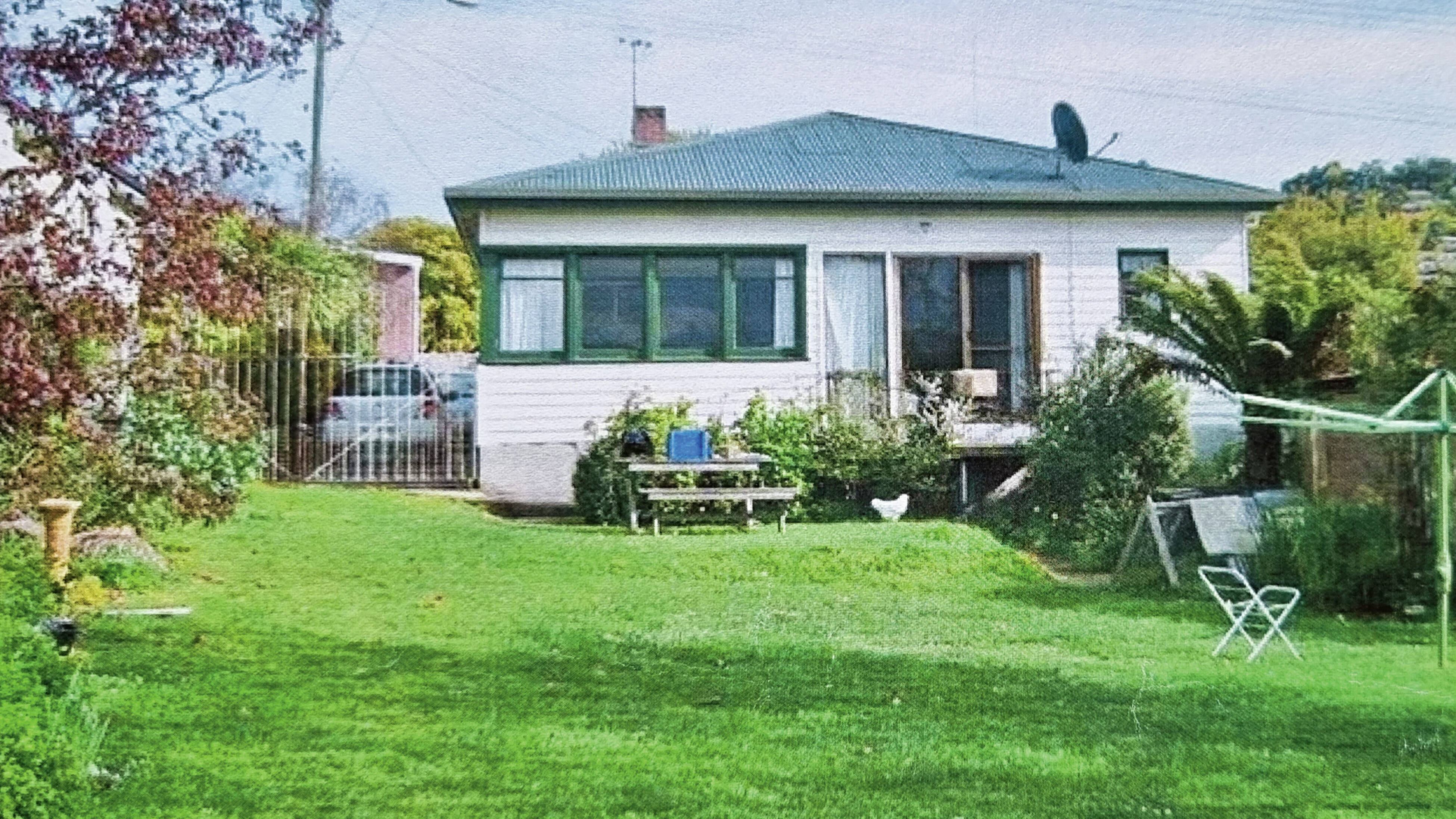 A white weatherboard home sits set back on a block of land at the top of a grassy slope with minimal landscaping.