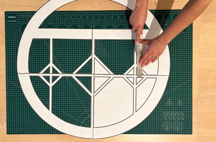 A pair of hands use a ruler and utility knife to cut out a paper window template on a green cutting mat]    