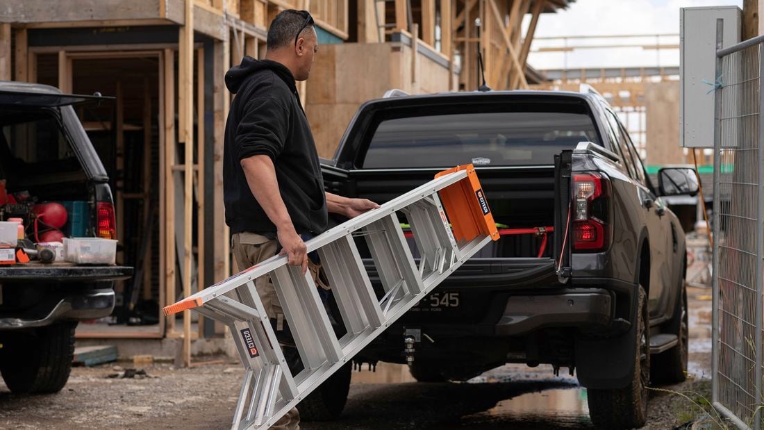 A tradie in work boots is putting a Citeco ladder into the back of a work ute on a job site.