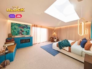 A modern bedroom featuring a large bed with white and brown bedding, colorful pillows, and a green throw blanket. The blue headboard wall has an arched design and pendant lights on either side. A wooden nightstand with decorative items sits next to the bed. The room includes a blue rug, beige curtains, and a rectangular skylight in the ceiling. 