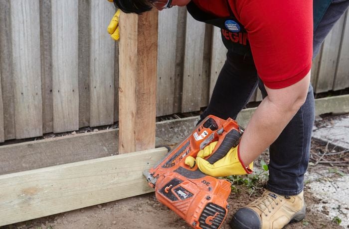A person attaching a plinth to a fence post using a nail gun