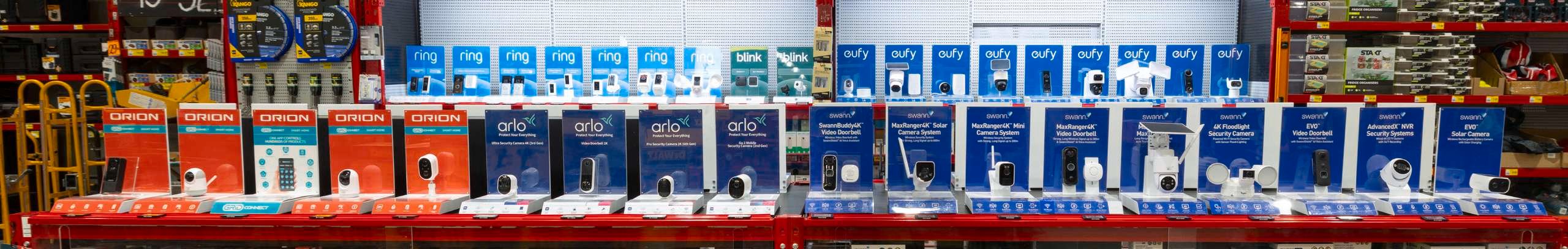 Close-up of a retail smart security display showcasing security cameras, video doorbells and home monitoring systems from brands including Ring, Eufy, Arlo, Swann and Orion. Products are arranged on red display shelving with boxed stock stored below.