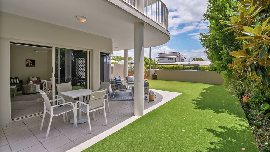 Ground-floor apartment patio with an outdoor dining setting and lounge area overlooking a private lawn bordered by established trees and gardens. Sliding doors connect the covered terrace to the interior living space.