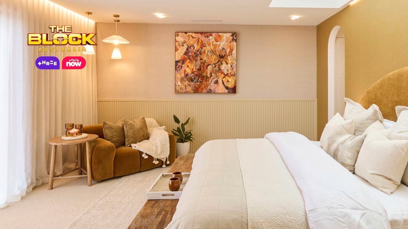 A stylish bedroom featuring a large bed with multiple pillows and a beige color palette. A wooden bench sits at the foot of the bed. To the left, there’s a cozy seating area with a brown sofa, cushions, and two small tables. Large windows with sheer white curtains let in natural light. The walls are decorated with artwork and lighting fixtures. 