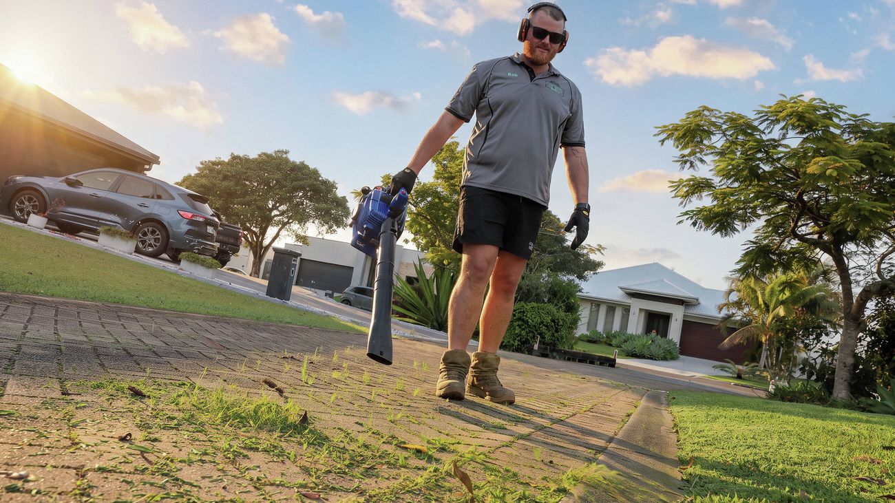 Person carrying a leaf blower while walking across a lawn in a suburban front yard.