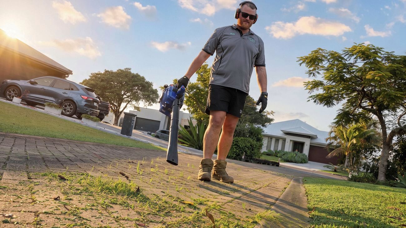 Person carrying a leaf blower while walking across a lawn in a suburban front yard.