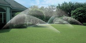 The backyard of a home showing lush green grass with five working irrigation sprinklers