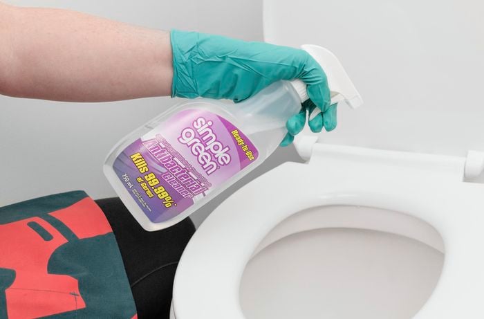 Person wearing a glove spraying toilet seat with liquid cleaner.
