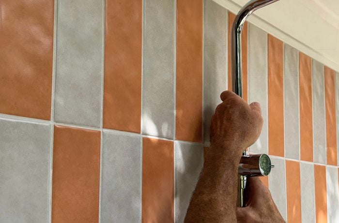 Hands tighten a chrome shower pipe on a wall of orange and grey rectangular tiles.