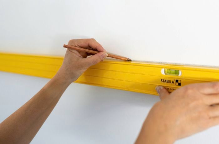 A person is using a spirit level and pencil to mark a straight line on the wall to set the height for a colour capping paint design. 