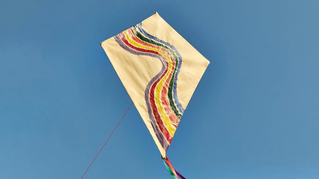 Beige diamond-shaped kite with retro rainbow stripes, attached to a red line, flying against a clear blue sky.