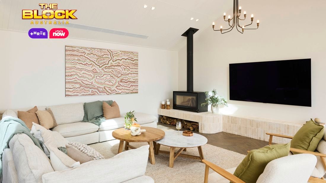A modern living room with high ceilings, featuring two chandeliers, a wall-mounted flat-screen TV, and a fireplace with logs stored below. The space includes a light-colored sectional sofa with cushions and throws, two armchairs with green cushions, and two round wooden coffee tables. Abstract artwork hangs above the sofa, and large windows bring in natural light. Decorative plants are placed throughout. 