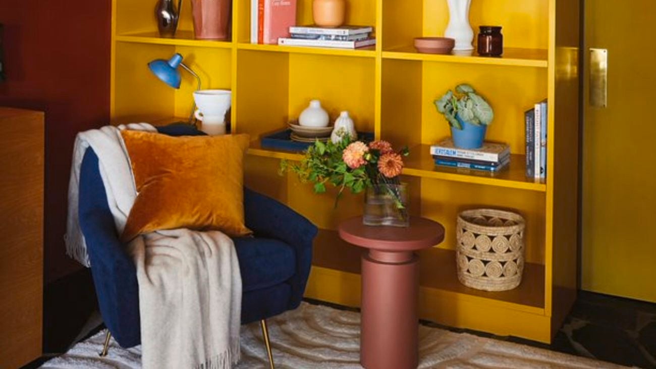 Yellow feature bookcase/shelving with blue velvet chair, with throw rug and cushions