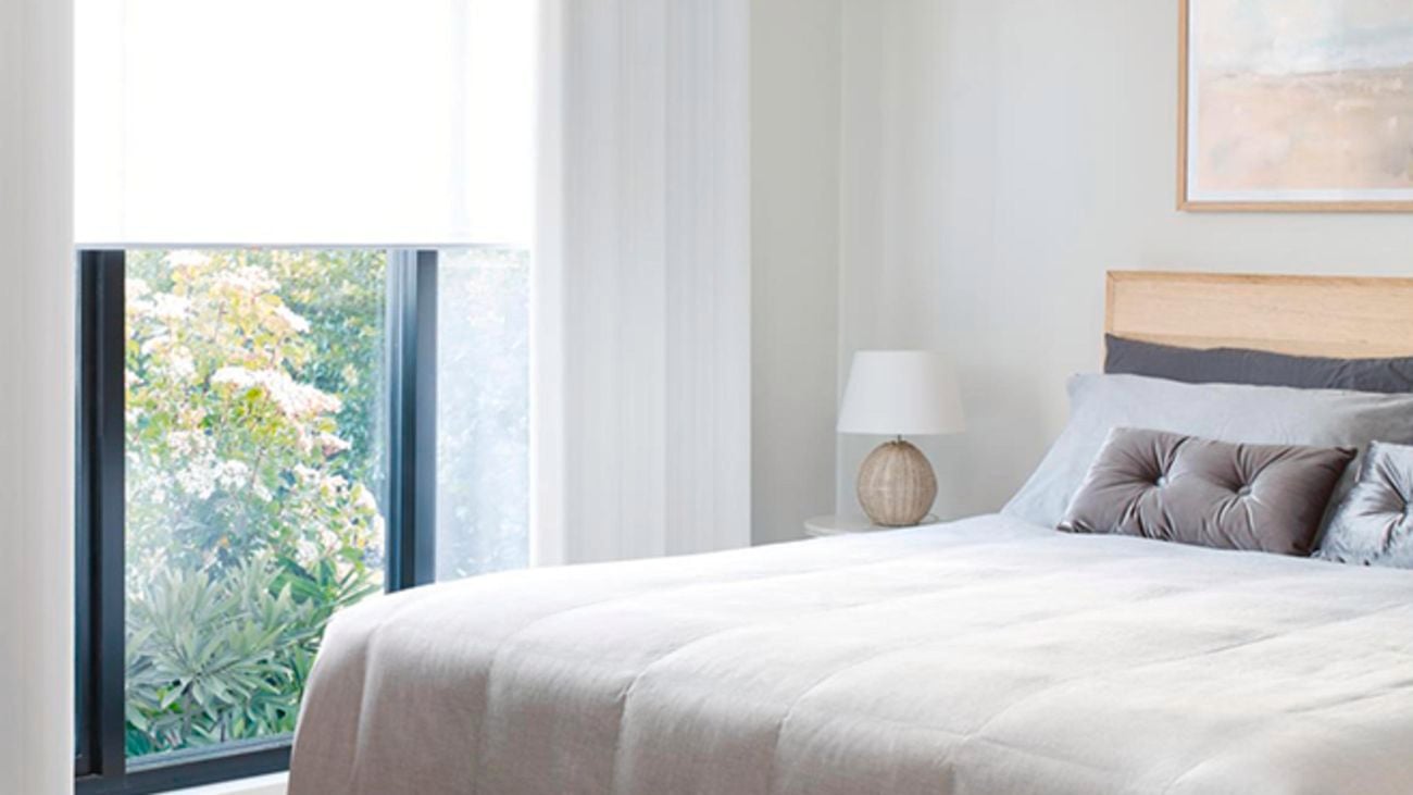 A bedroom with a bed, a window, roller blinds and white sheer curtains