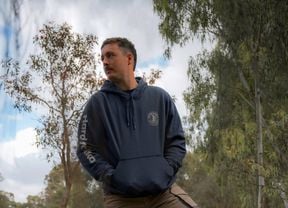 Person standing outdoors in a wooded area with one foot on a tree stump, wearing a hoodie and work pants, with a chainsaw on the ground nearby.