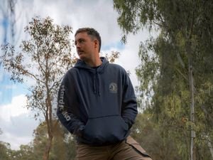 Person standing outdoors in a wooded area with one foot on a tree stump, wearing a hoodie and work pants, with a chainsaw on the ground nearby.