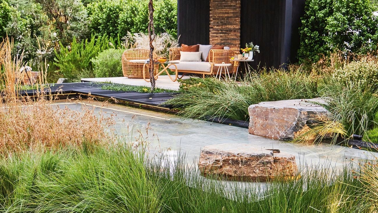 A garden with a hardscaped area featuring outdoor furniture next to a water feature surrounded by metal platforms and plantings of rush and kangaroo grass with a large rectangular boulder.