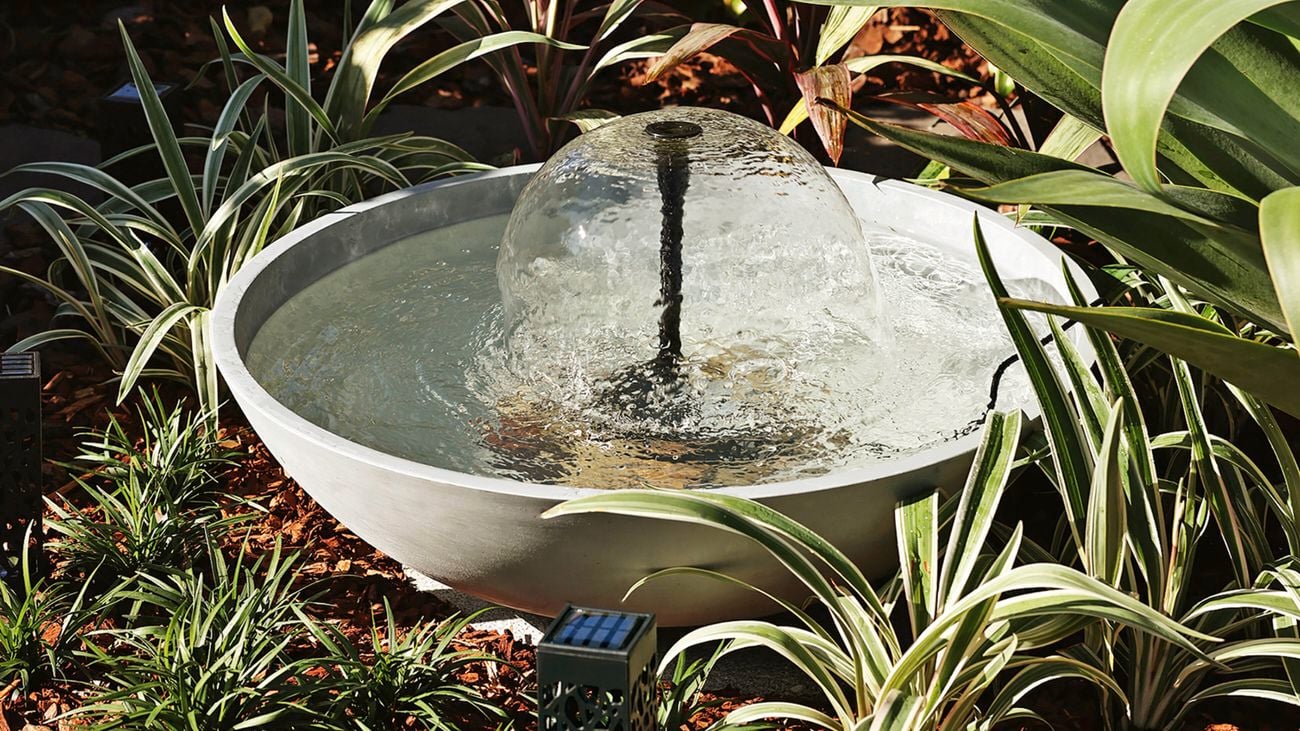 A solar-powered planter bowl water feature with a central bubbler sits on a garden bed of mulch and green plants.