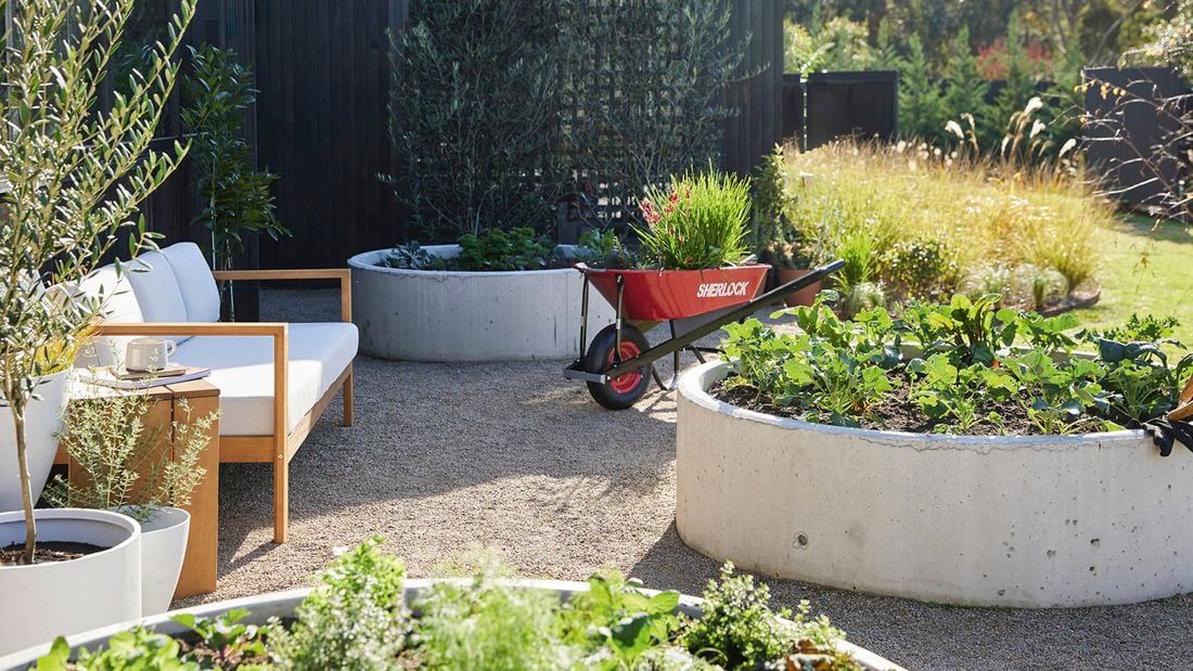 Exterior of a black timber modern farmhouse with an outdoor area featuring a red wheelbarrow, raised garden beds, potted plants, and a white outdoor sofa on gravel. Trees, lawn and greenery are visible in the background.