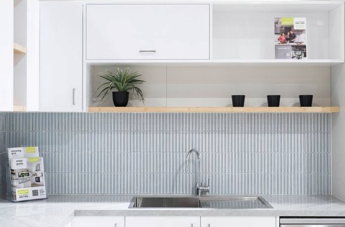 A kitchen with a grey bench top, mixer and sink in it, shelving with white cupboards in them attached to the wall.