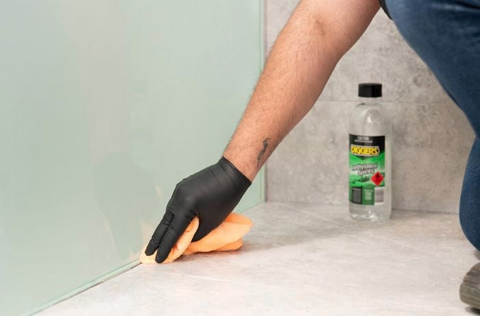  A  person wearing gloves is using a clean cloth and methylated spirits to prepare bathroom surfaces before silicone application.