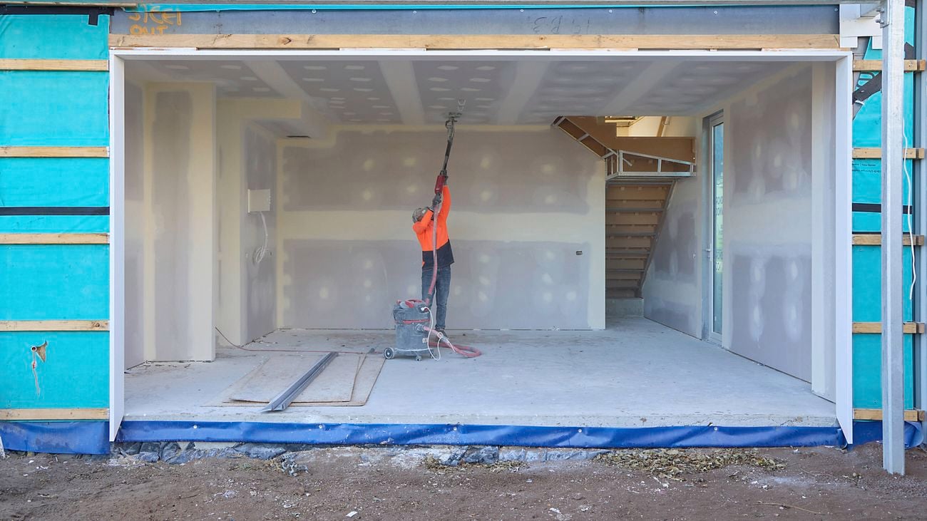 A tradie in orange hi-vis uses a vacuum on ceiling fittings at the mid-way point on a construction site.