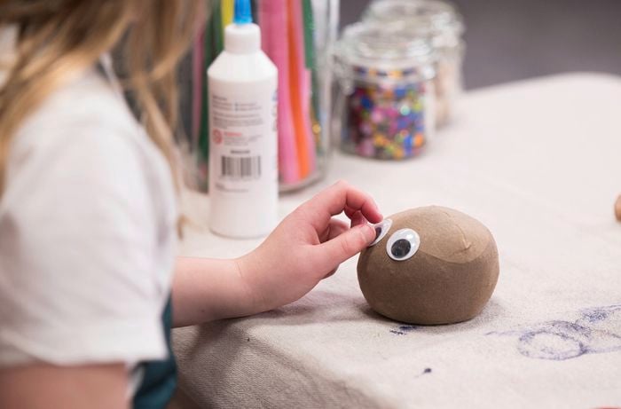 A girl is applying googly eyes to a grass head face using craft glue. 