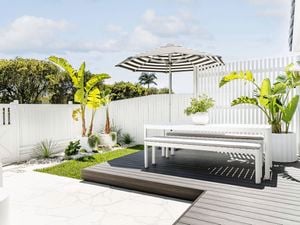 A backyard design setting with a black and white striped sun umbrella, white outdoor setting and fence, plus greenery in white pots